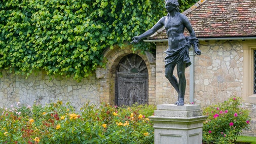 Apollo sculpture in the Rose Garden at Anglesey Abbey. The statue is on a raised stand, arm outstretched and surrounded by yellow roses.